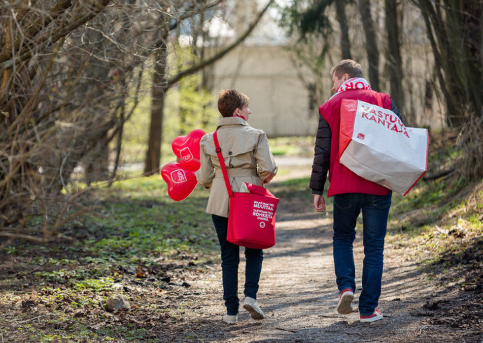 Nainen ja mies metsäpolulla, naisella SDP ilmapalloja ja laukku, missä teksti Maailmaa voi muuttaa, tarvitaan vaan rohkeita ihmisiä, Miehellä laukku missä teksti vastuunkantaja